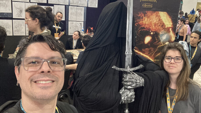 A imagem mostra duas pessoas posando para uma selfie com um personagem encapuzado que segura uma espada, em um ambiente de feira de quadrinhos ou evento cultural. (Legendado por IA)