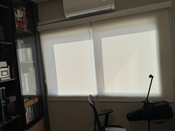 A imagem mostra um ambiente interno com uma cadeira de escritório, um ar-condicionado no teto e janelas cobertas por cortinas, ao lado de uma estante cheia de jogos. (Legendado por IA)