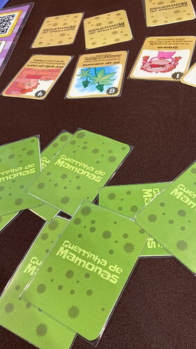 A imagem mostra cartas de um jogo chamado "Guerrinha de Mamonas" dispostas sobre uma mesa, com algumas cartas visíveis e focadas em temas cômicos e divertidos. (Legendado por IA)