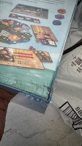 A imagem mostra a borda de uma caixa de jogo de tabuleiro, com detalhes gráficos visíveis na parte traseira e um pacote ao lado. (Captioned by AI)