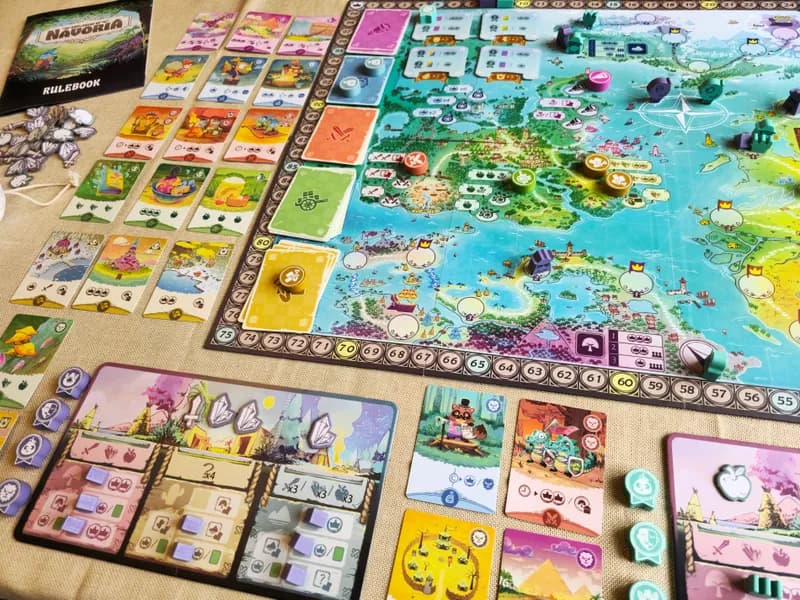 A imagem mostra um jogo de tabuleiro chamado "Nadoria", com o tabuleiro central, várias cartas e peças dispostas ao redor. (Captioned by AI)